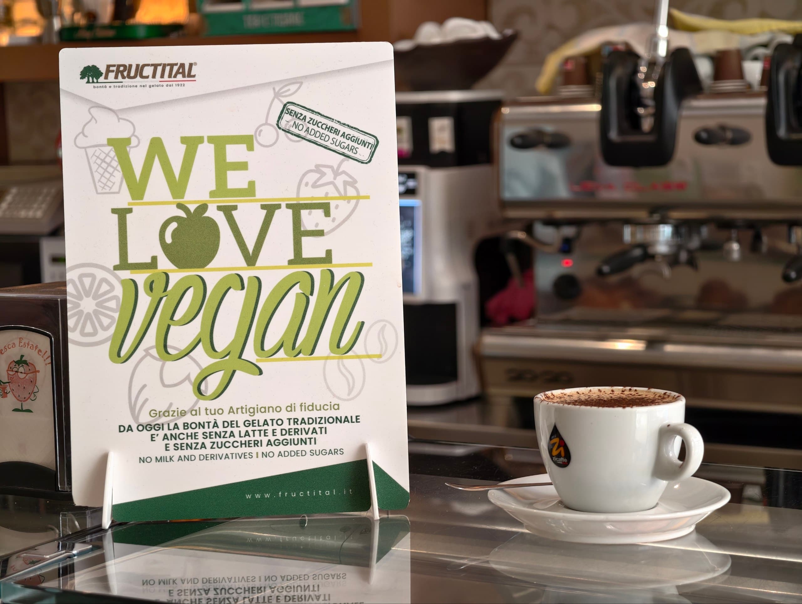 Cartello "We Love Vegan" e tazzina di caffè su un bancone di bar con macchina del caffè sullo sfondo.
