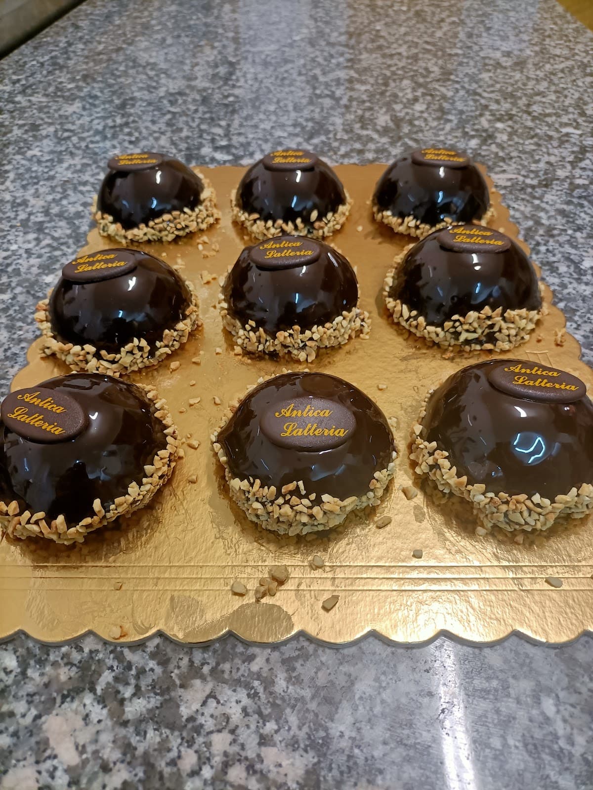 Nove dolci monoporzione a cupola con glassa al cioccolato e granella di nocciole, presentati su un vassoio dorato.