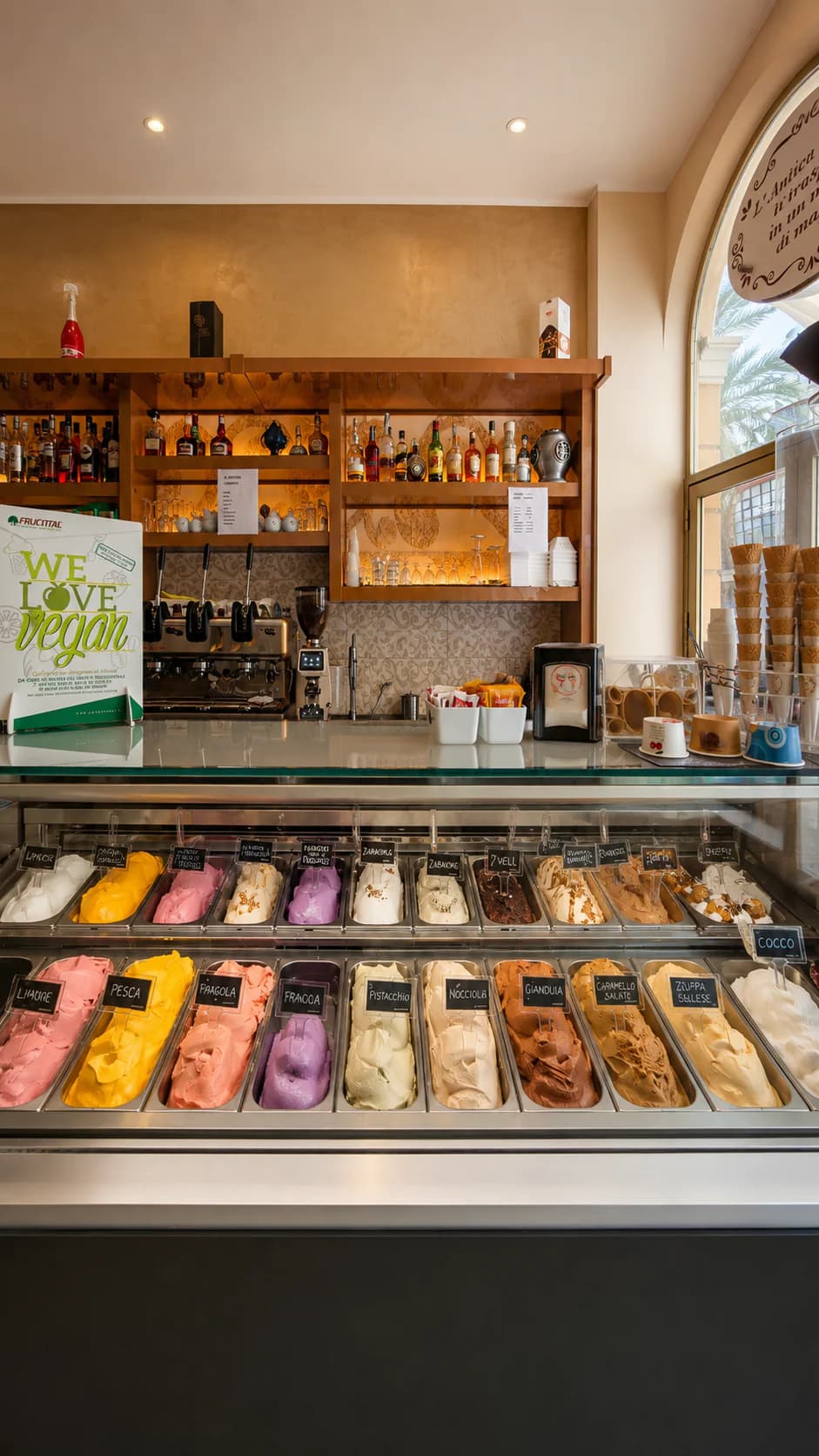Vetrina di gelateria con numerosi gusti di gelato, macchine per il caffè e bottiglie di liquori sullo sfondo.