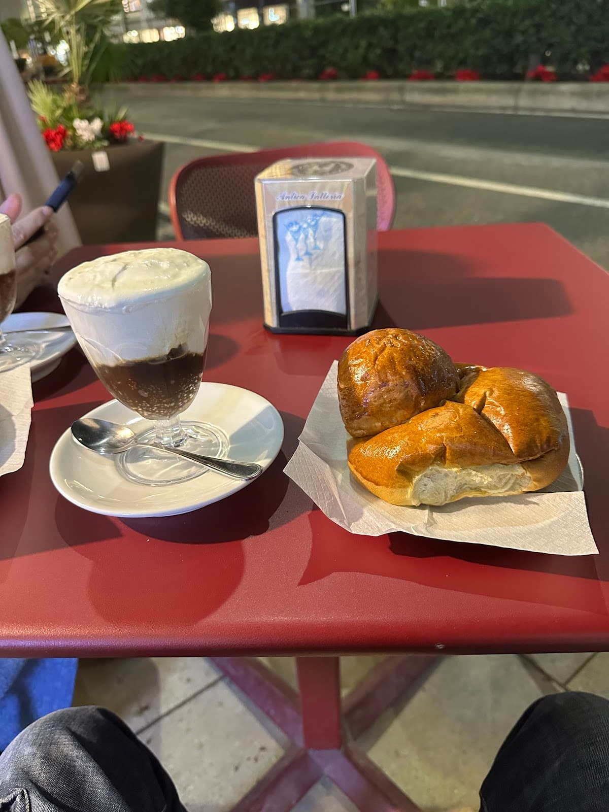 Un bicchiere di caffè con abbondante panna e un cornetto su un tavolo rosso, con un portatovaglioli in evidenza.