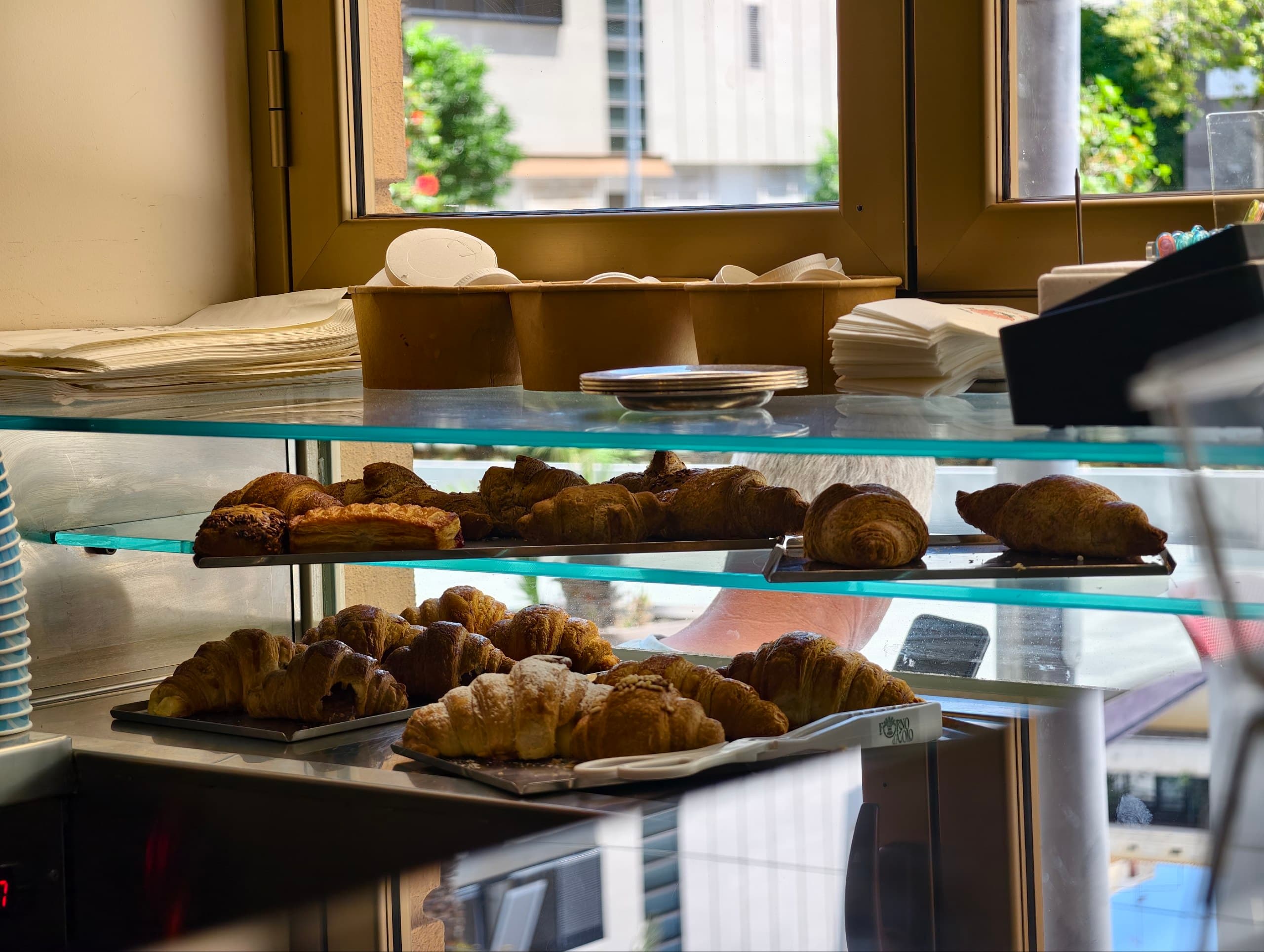 Croissant e dolci da forno esposti in una vetrina di un bar, con vista esterna sfocata.