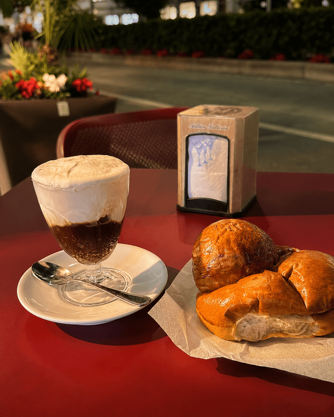 Un bicchiere di caffè con panna e due brioche su un tavolo rosso, con un portatovaglioli e fiori sullo sfondo.