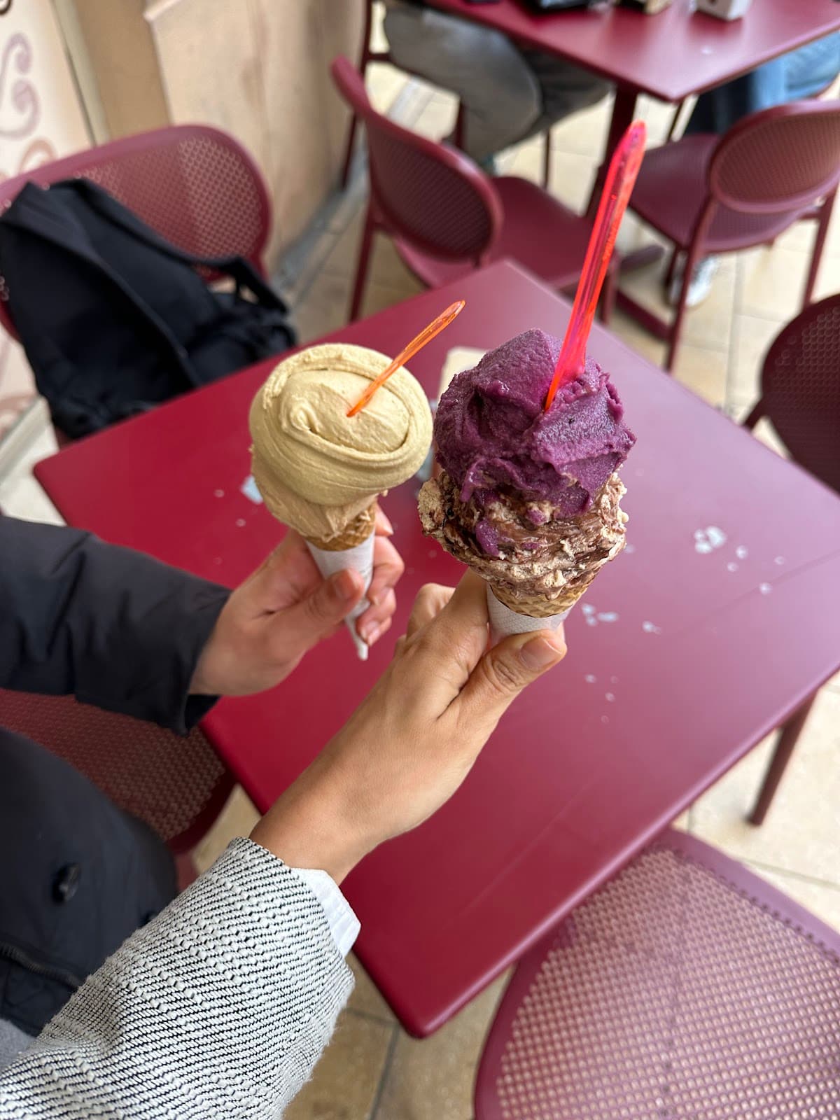 Due mani che tengono coni gelato artigianale, uno color crema e uno viola, su un tavolo rosso.